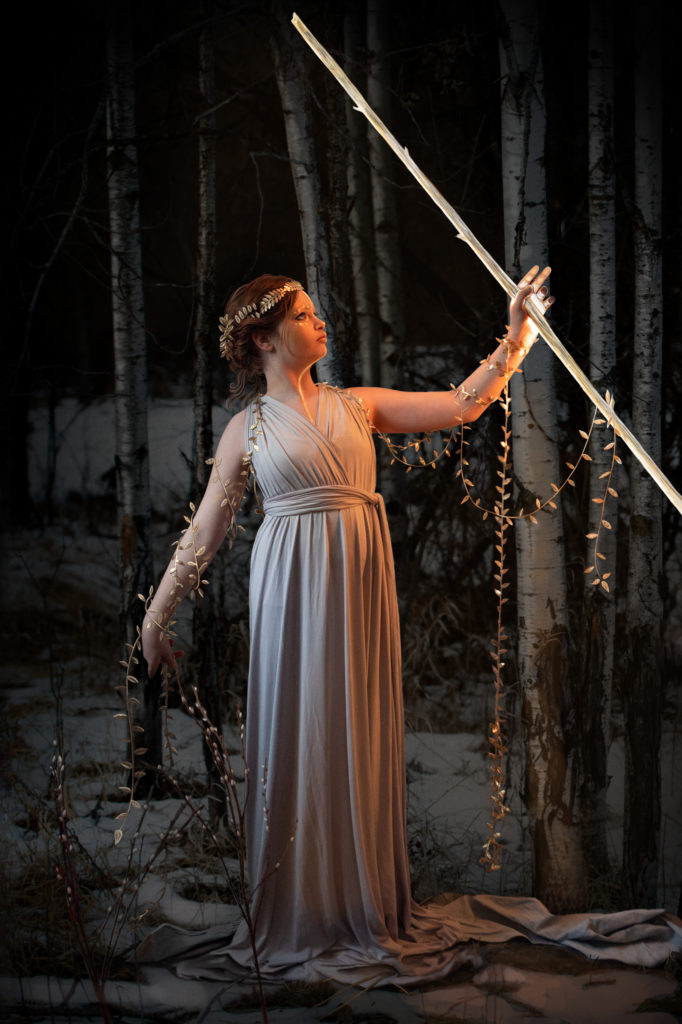 Young woman in simple draped dress, standing in a grove of trees, with gold leaves on vines around her arms. She has gold paint down her face and sternum, and holds a glowing staff in the air.